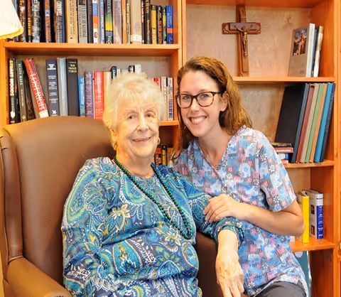 An elderly woman sitting in a brown armchair next to a younger woman in scrubs, both smiling. They are in a room with wooden bookshelves filled with books and a crucifix on the wall behind them.