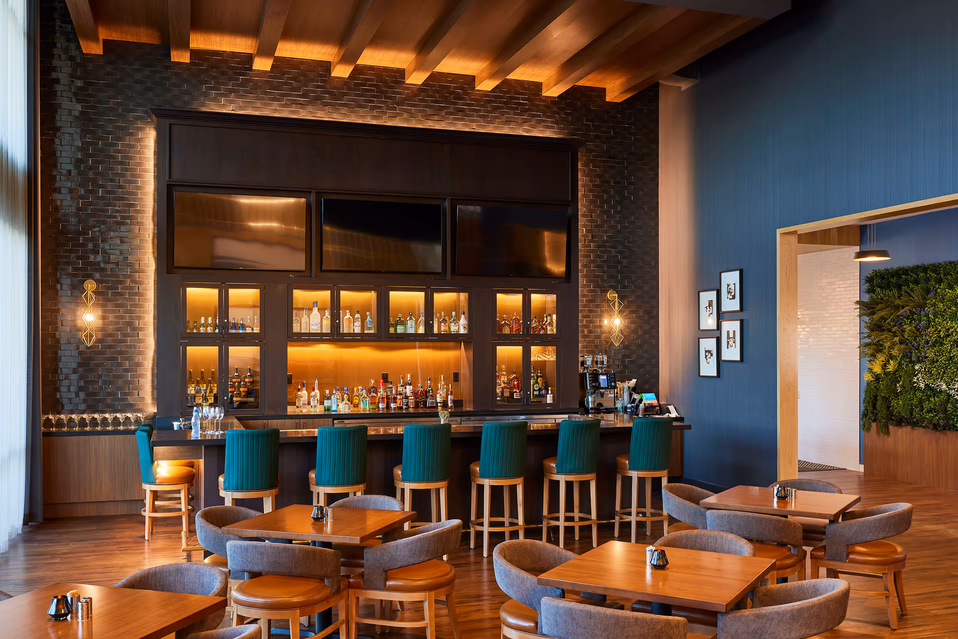Interior view of a stylish bar area with a long counter lined with green cushioned bar stools. Behind the bar, shelves display various bottles of liquor illuminated by warm lighting. The room features wooden floors, a dark brick wall, and a ceiling with wooden beams. Several wooden tables with cushioned chairs are arranged in front of the bar. The right side of the image shows a blue wall with framed artwork and a doorway leading to another room with a green plant wall.
