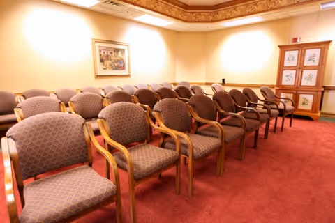 Rows of upholstered wooden chairs arranged in a carpeted meeting room with framed artwork and a decorative cabinet.