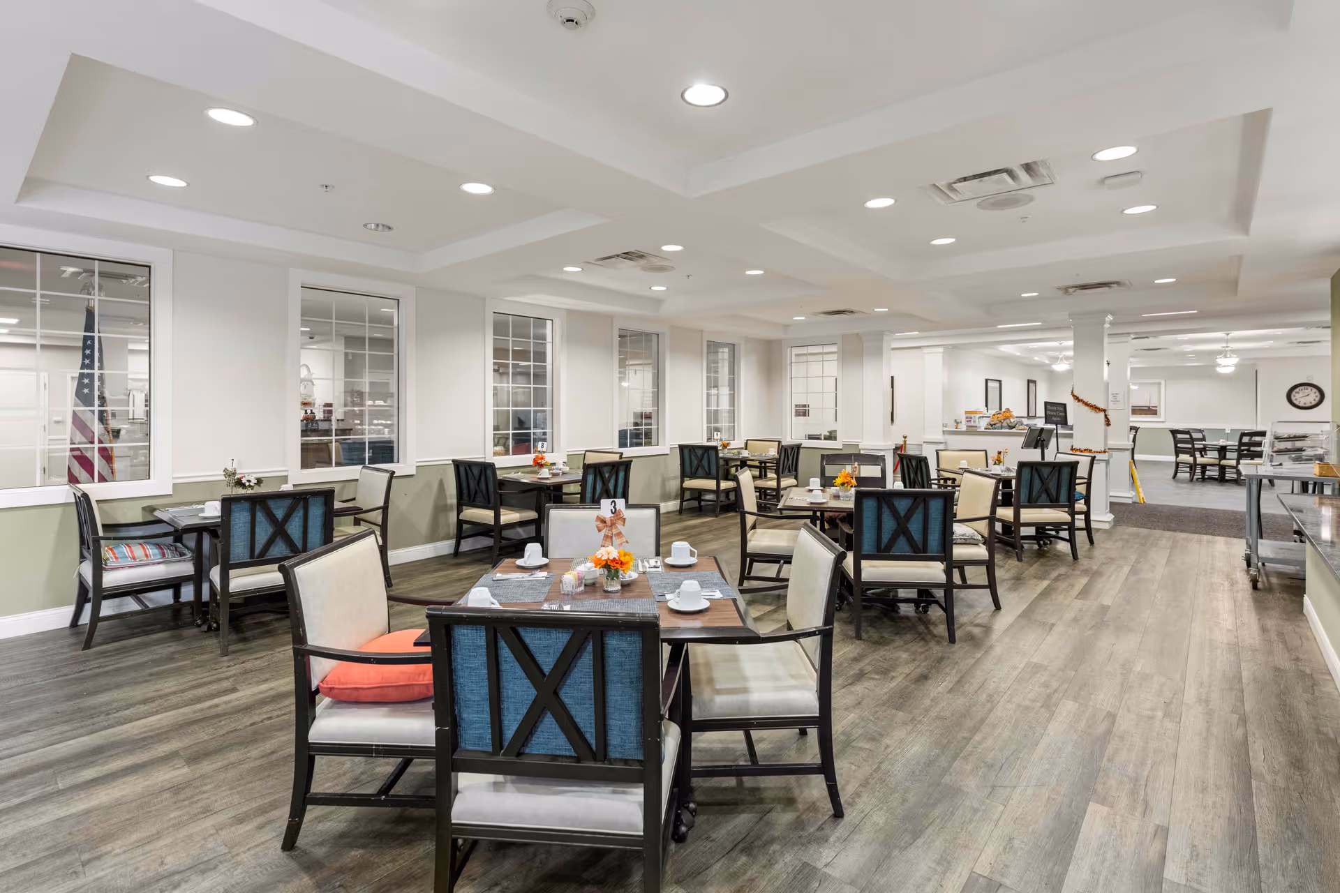 A bright and spacious dining room in a senior living facility with multiple tables and chairs arranged neatly. Each table is set with white cups, plates, and small floral centerpieces. The room has large windows, recessed lighting, and wood-style flooring. The walls are painted white with a light green accent below the windows.