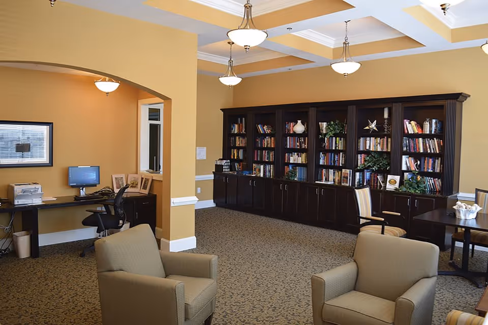 A cozy interior room with beige walls and carpeted floor featuring two beige armchairs in the foreground. In the background, there is a large dark wooden bookshelf filled with books and decorative items. To the left, there is a desk with a computer, printer, and office chair under an archway. The ceiling has recessed lighting fixtures.