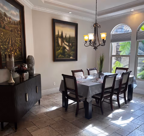 Well-lit dining room with a rectangular table set for six under a chandelier, a sideboard with decor, framed paintings, and arched windows letting in sunlight.