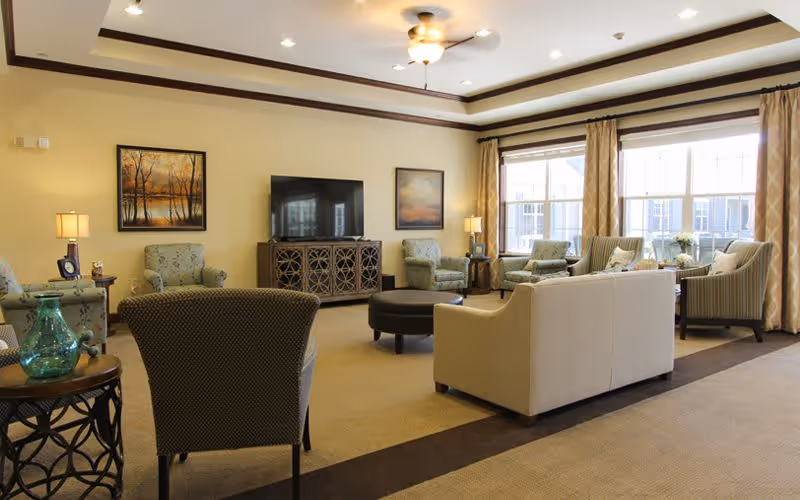 A spacious living room with multiple upholstered armchairs and sofas arranged around a central ottoman. The room features large windows with curtains allowing natural light to fill the space, a flat-screen TV on a decorative cabinet, two framed paintings on the wall, and several table lamps providing warm lighting.