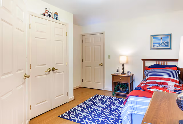 A bedroom with a single bed dressed in red, white, and blue striped bedding. There is a wooden nightstand with a lamp, a small clock, and a decorative item. A framed picture of a lighthouse hangs on the wall above the bed. The floor has a blue patterned rug, and there are three closed doors visible, two on the left and one straight ahead.