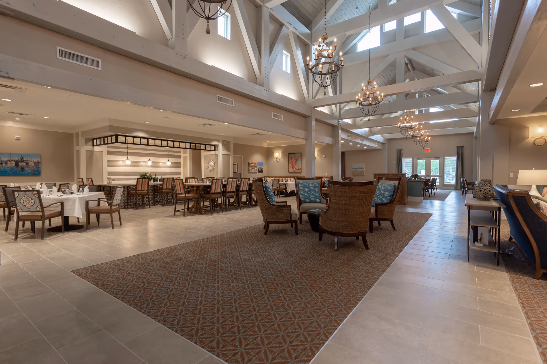 Spacious interior of a senior living facility with high vaulted ceilings and exposed beams. The room features a seating area with wicker chairs and blue cushions on a patterned carpet, multiple dining tables with chairs, and a bar area with stools. Large windows and glass doors allow natural light to fill the space, and several chandeliers hang from the ceiling.