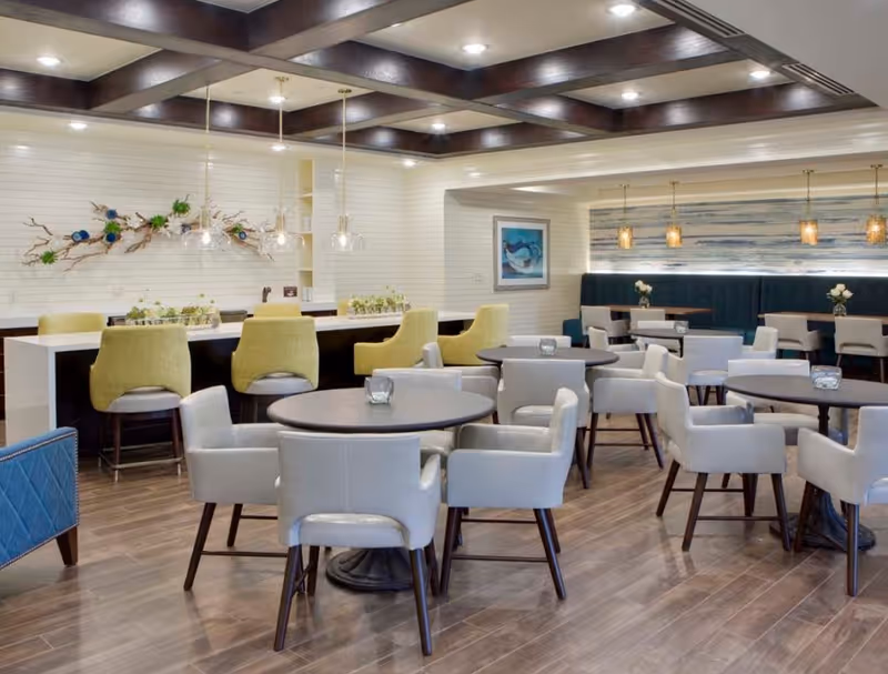 Open dining area with round tables and upholstered chairs, a long counter with yellow bar stools, decorative wall art, and coffered ceiling lighting.