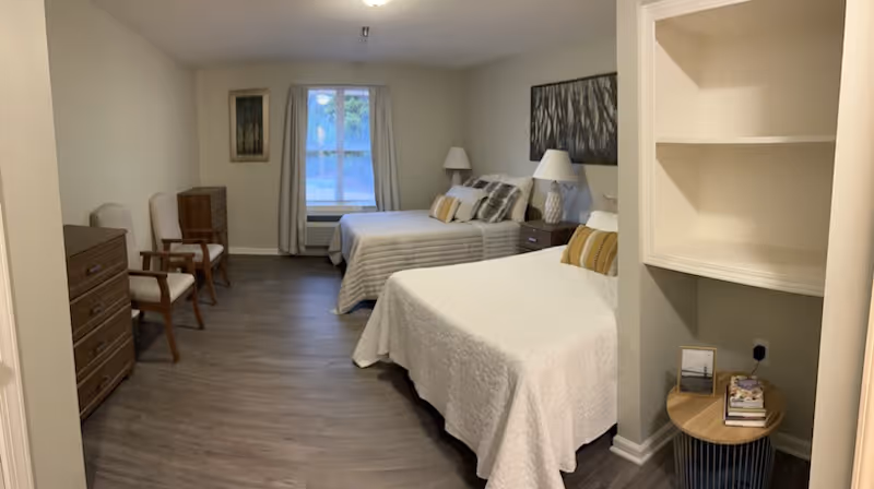 A senior living facility bedroom with two beds covered in white and striped bedspreads, each with pillows. There are two bedside tables with lamps, two wooden chairs, a dresser, a window with curtains, and a small round table with books and a framed photo. The room has wooden flooring and neutral-colored walls.