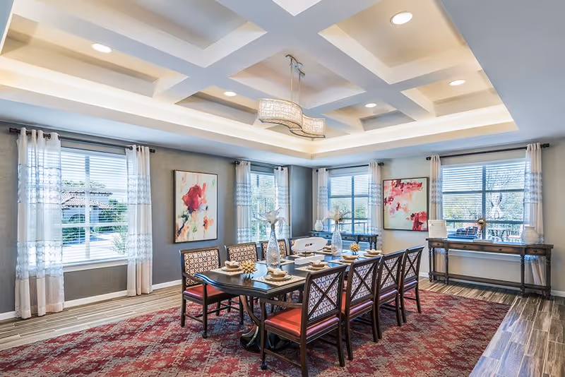 A bright and elegant dining room with a long dark wooden table set for eight people. The chairs have patterned backs and red cushions. The room features large windows with sheer white curtains, colorful abstract paintings on the walls, a decorative ceiling with recessed lighting, and a red patterned carpet covering the floor. A side table with decorative items is placed against one wall.