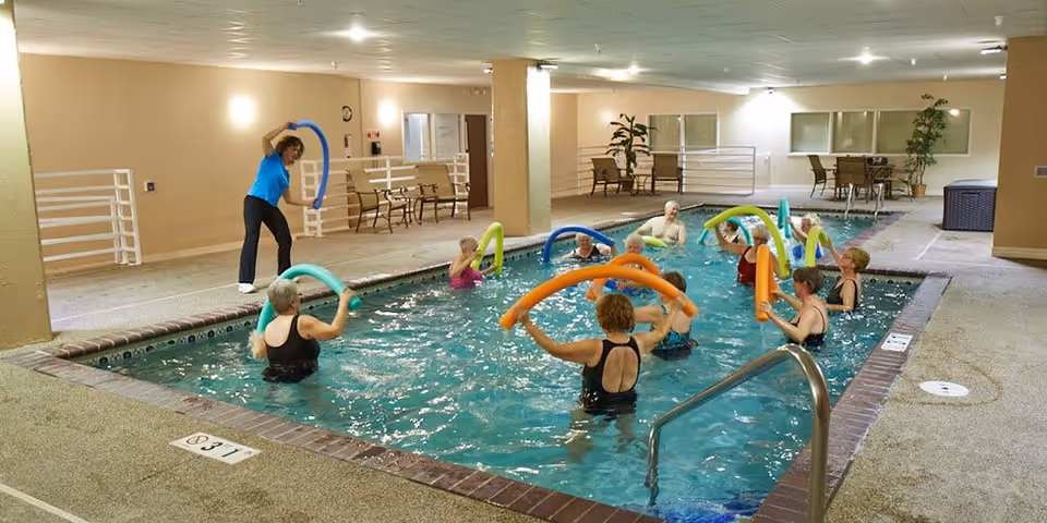 A group of elderly people participating in a water exercise class in an indoor swimming pool, following the instructor who is standing poolside holding a blue pool noodle. The pool area has beige walls, benches, and some plants in the background.