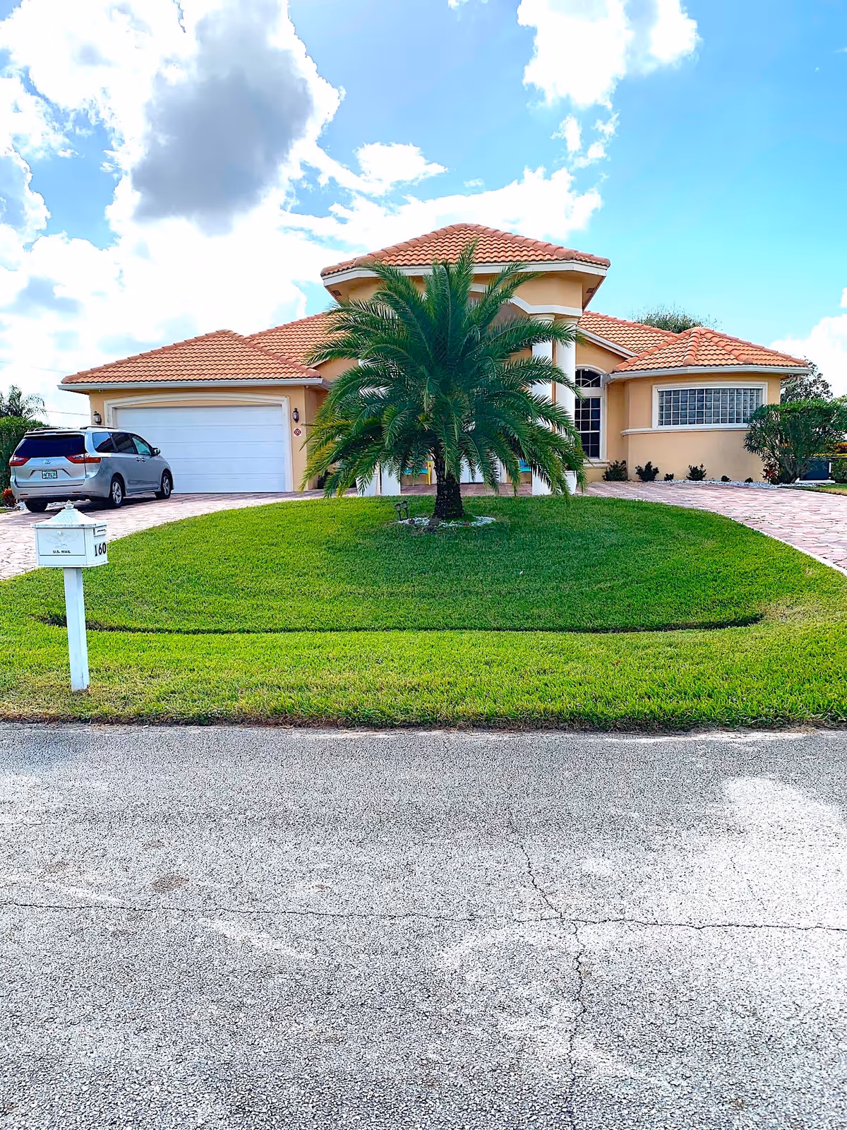 Front view of a Mediterranean-style house with a tiled roof, a palm tree centered on the lawn, driveway, garage and mailbox.