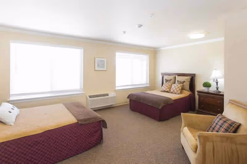 A bright bedroom with two beds, each with a brown and beige bedspread and decorative pillows. There are two large windows with white blinds, a wooden nightstand with a lamp and a small plant, and a beige armchair with a plaid cushion. The walls are light-colored and the floor is carpeted.