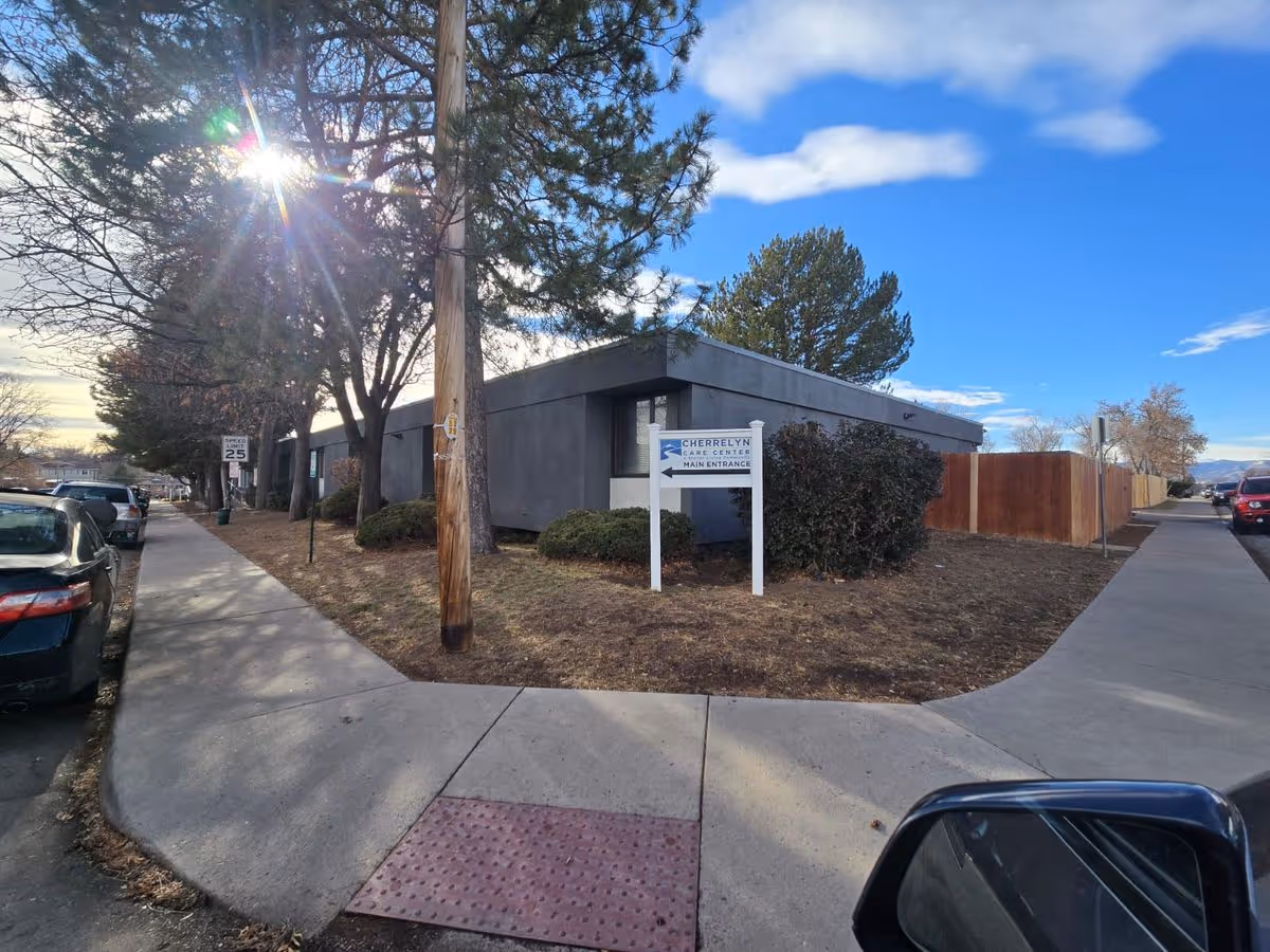 Street-level view of the Cherrelyn Care Center single-story building with a main entrance sign, sidewalk, parked cars and trees under a blue sky.