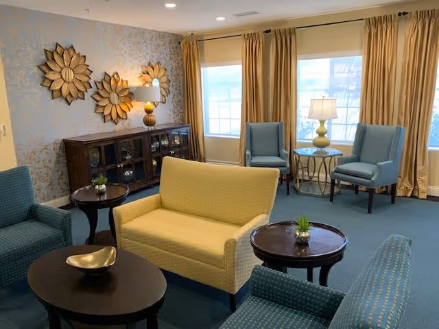 A cozy living room area with a yellow loveseat, two blue armchairs, and two small round wooden tables with decorative plants. There is a wooden cabinet against the wall with three large decorative sunburst mirrors above it. Two windows with beige curtains allow natural light into the room, and a table lamp is placed on a small round table between two blue chairs.