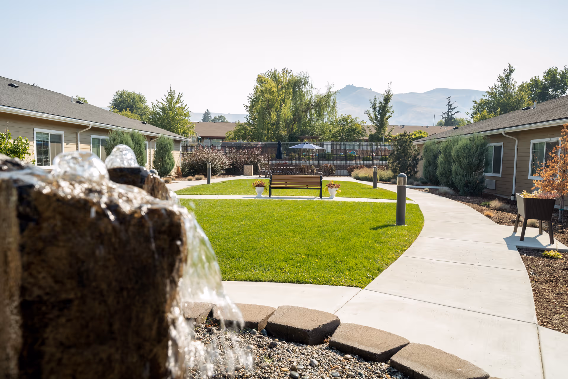 Outdoor courtyard area at Ciel of Wenatchee featuring a water fountain in the foreground, a curved concrete walkway, green lawn, benches, potted plants, and single-story buildings with mountains visible in the background under a clear sky.