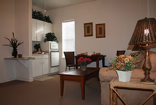 A cozy living area with a beige sofa, a wooden coffee table with a flower pot, a dining table with chairs, and a small kitchen area with white cabinets and appliances. The room is decorated with plants, framed pictures on the wall, and a table lamp.