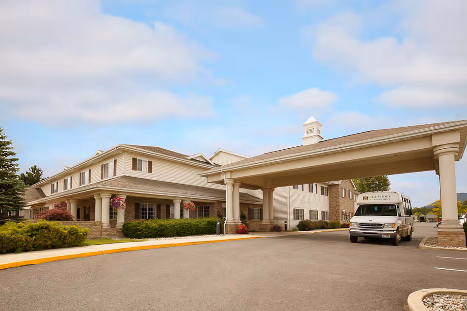 Front exterior of The Bridge at Sandpoint senior living building with a covered porte-cochere and a shuttle van parked nearby.