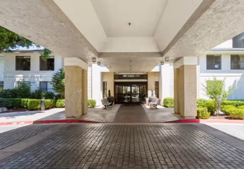 Covered porte-cochere entrance to a residential building with benches, columns, and landscaped shrubs.