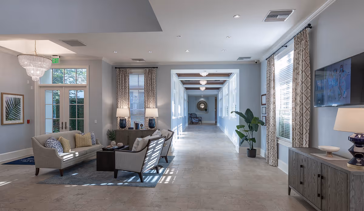 A spacious, well-lit interior hallway with light gray walls and tiled flooring. To the left, there is a seating area with a gray sofa, two armchairs, a dark coffee table, and decorative pillows. Two table lamps sit on a console table against the wall, flanked by windows with patterned curtains. A chandelier hangs near the entrance door with glass panels. On the right side, there is a large window with matching curtains, a flat-screen TV mounted on the wall, and a wooden cabinet with a lamp and decorative items. The hallway extends into the distance with additional seating and a round mirror on the far wall.