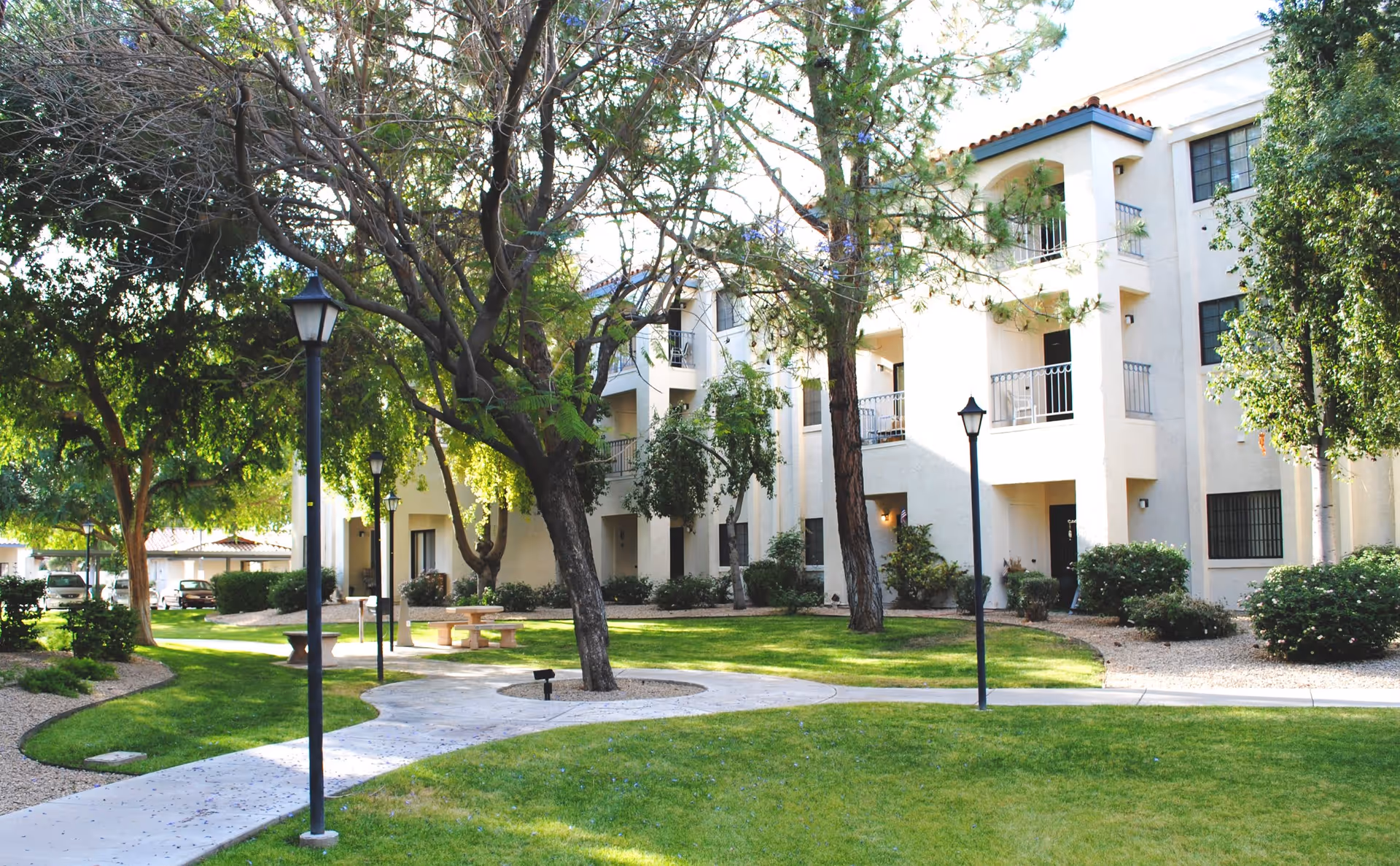 Outdoor view of a senior living facility courtyard with green grass, trees, lamp posts, and a paved walkway. The building has multiple balconies and is painted in a light color with a tiled roof.