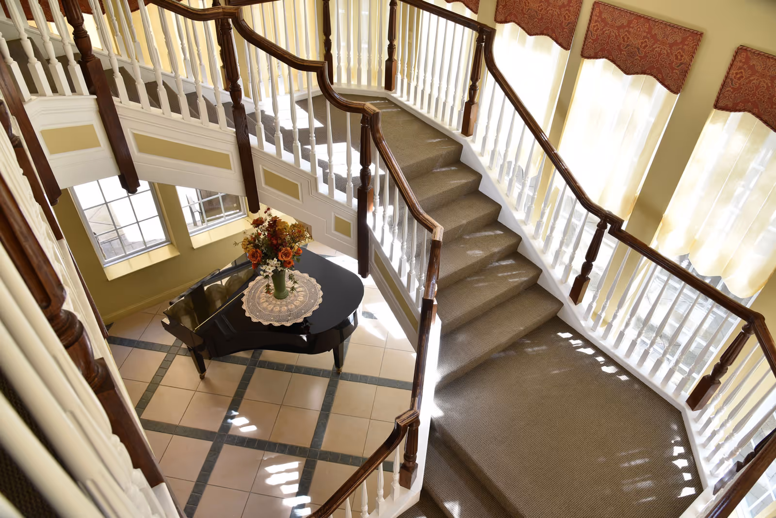 View from above of a carpeted staircase with white railings and dark wooden handrails, leading down to a tiled floor area with a black grand piano decorated with a lace doily and a vase of flowers. Large windows with sheer curtains allow natural light to fill the space.