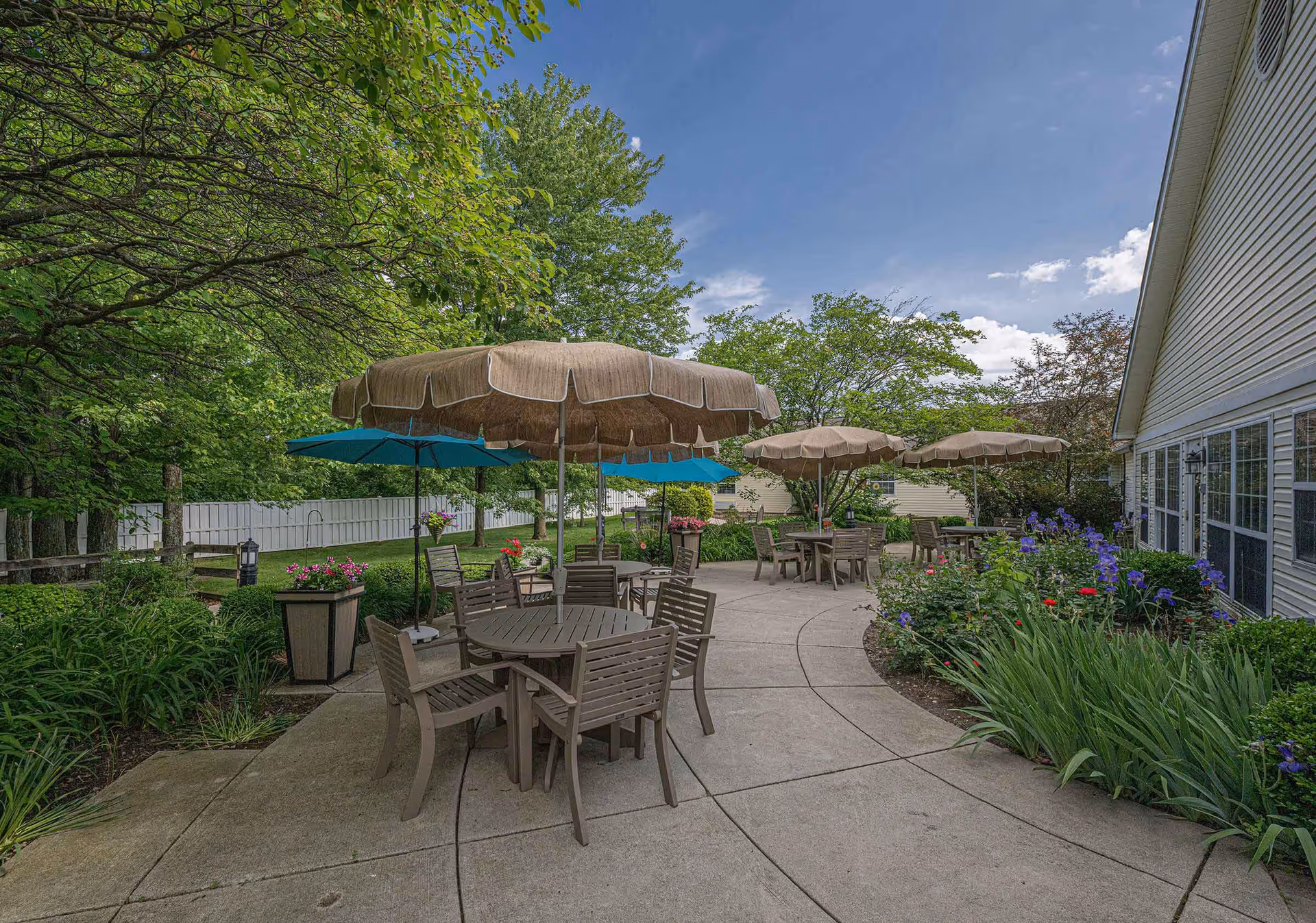 Outdoor patio area with multiple round tables and chairs under large beige and blue umbrellas. The patio is surrounded by green trees, bushes, and colorful flowers, with a building visible on the right side under a partly cloudy blue sky.