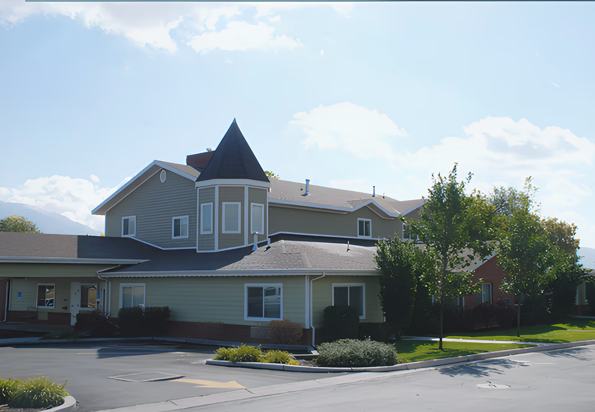Exterior view of The Lodge at Jordan River, a two-story building with light green siding and a distinctive turret with a pointed roof. The building is surrounded by trees, shrubs, and a paved driveway under a partly cloudy sky.