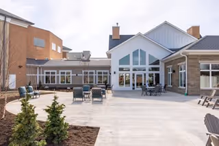 Outdoor patio area of a senior living facility with several tables and chairs arranged on a concrete surface, surrounded by buildings with large windows and a few small evergreen shrubs planted in a mulched area.