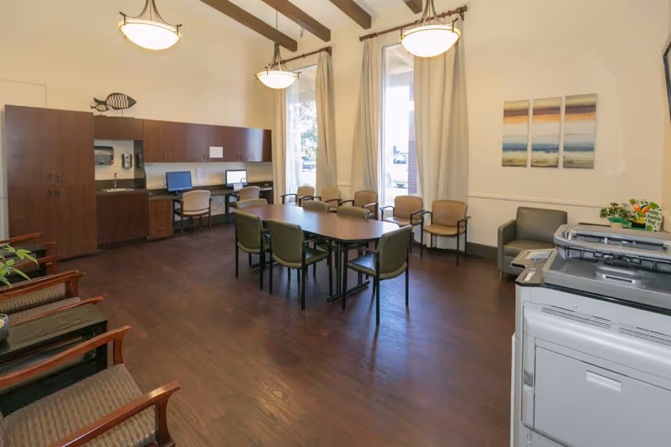 A spacious room with a long table surrounded by chairs in the center, two computer workstations with chairs against the back wall, wooden cabinets, large windows with curtains, a printer/copier machine in the foreground, and a few armchairs along the walls. The room has wooden flooring and ceiling beams with hanging lights.