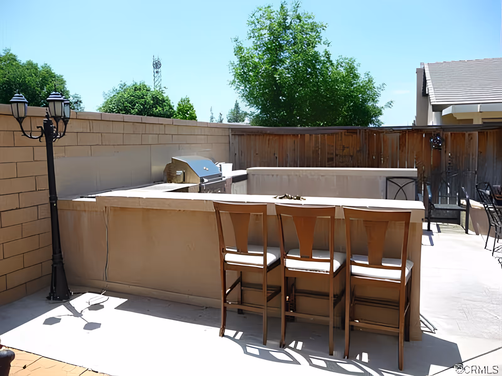 Outdoor patio area with a built-in bar and grill, three barstools, a lamp post and wooden fence under a clear sky.