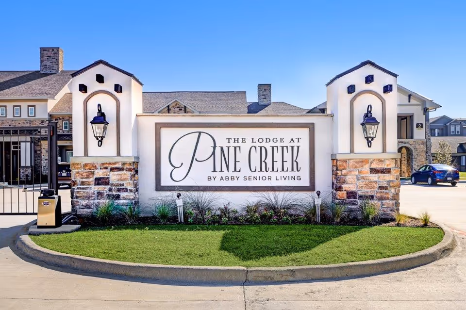 Entrance sign for The Lodge at Pine Creek by Abby Senior Living, featuring stone pillars with lanterns on either side and a well-maintained grassy area in front, with part of the building and a parked car visible in the background under a clear blue sky.