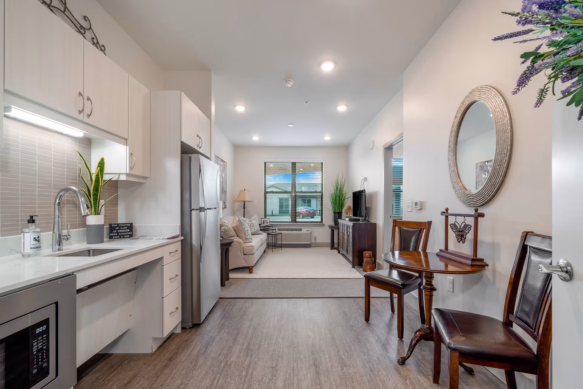Open-plan apartment interior showing a small kitchen with stainless appliances and a dining area leading to a furnished living room.