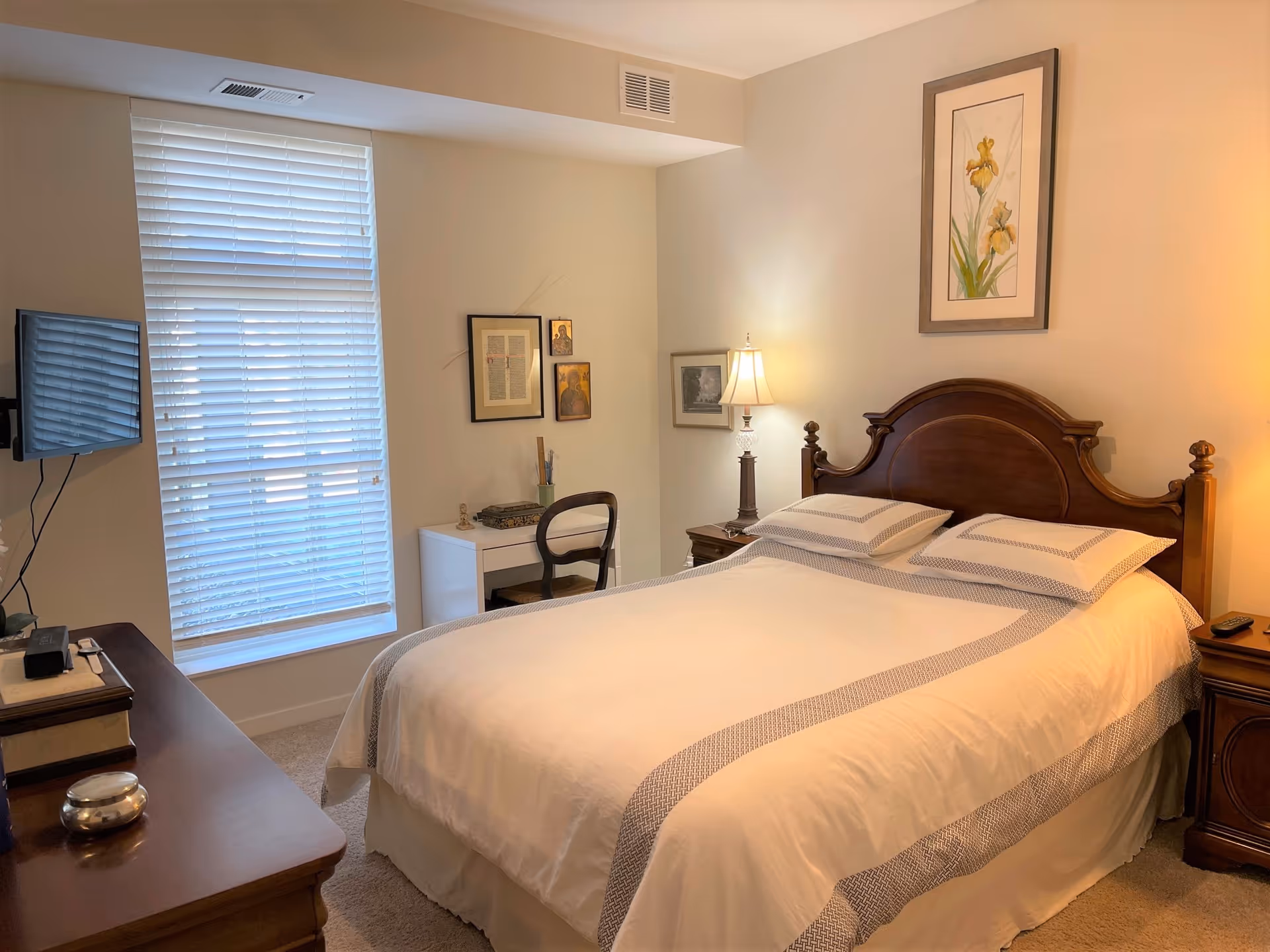Well-lit bedroom with a made double bed, wooden headboard, bedside tables and lamp, a small desk, TV, and a window with blinds.