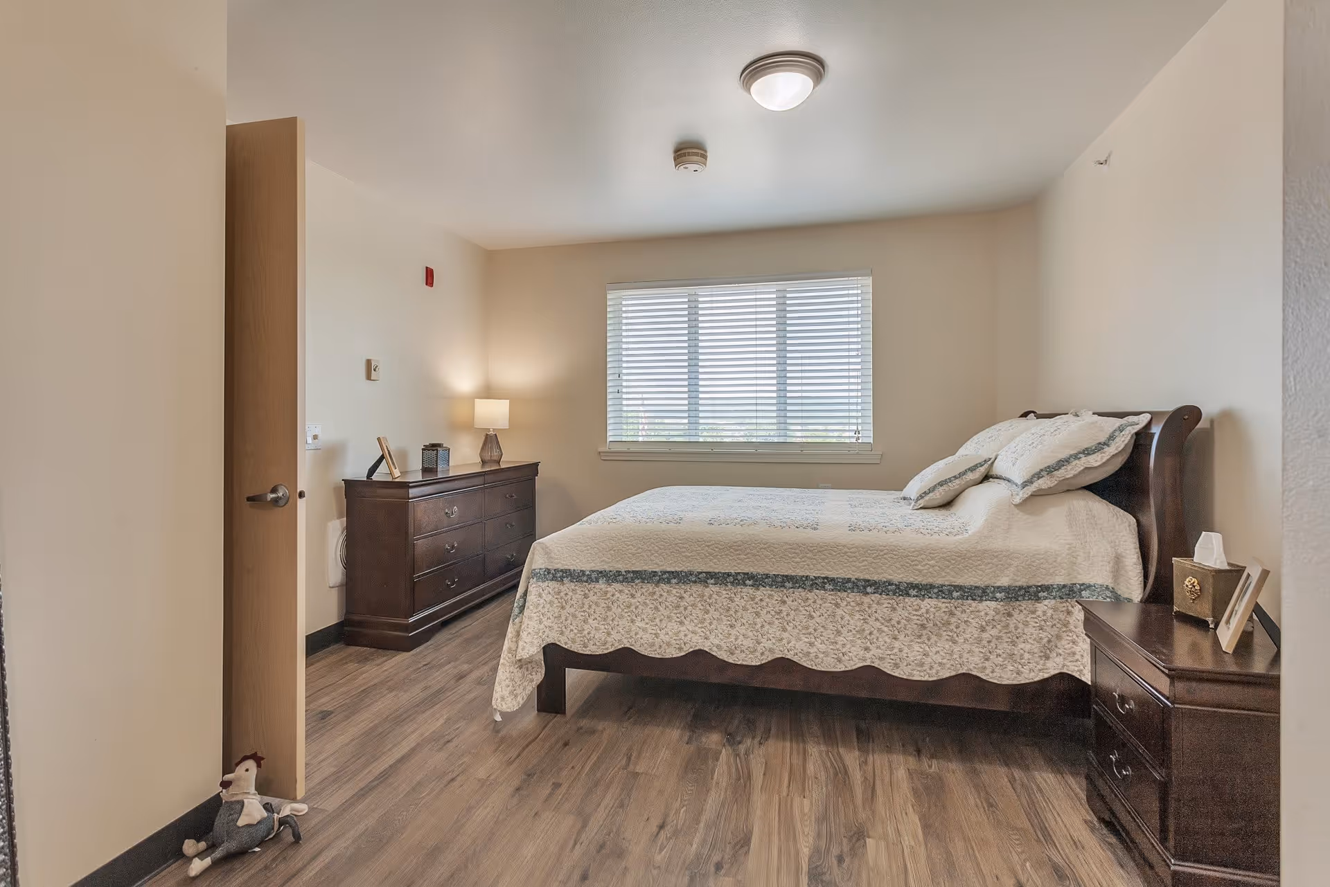 A bedroom in a senior living facility with a wooden bed covered with a white and blue quilt, two pillows, a wooden nightstand with a tissue box and picture frame, a wooden dresser with a lamp and decorative items, a window with blinds, and a plush toy on the floor near the door.