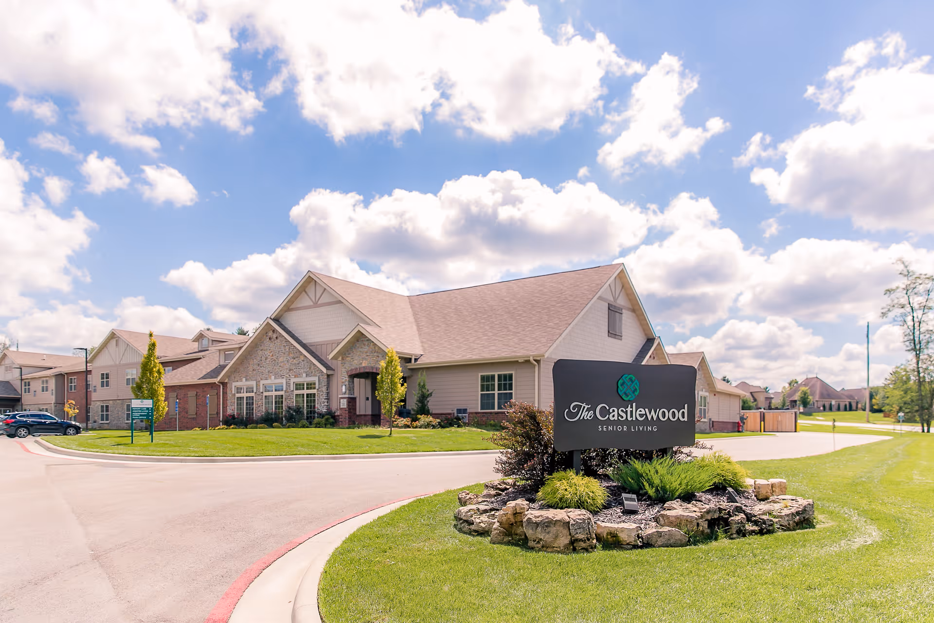 Front exterior of The Castlewood Senior Living building with its entrance sign on a landscaped roundabout under a partly cloudy sky.