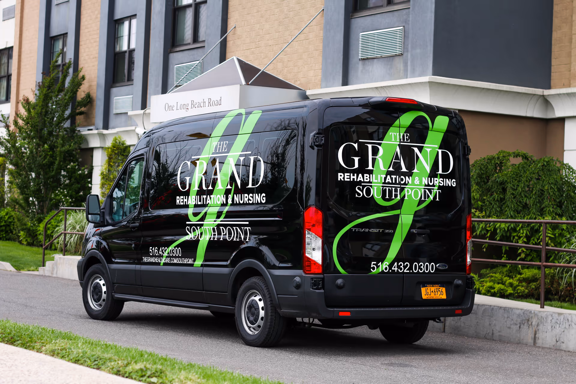 A black van parked outside a building with signage for The Grand Rehabilitation & Nursing at South Point. The van displays the facility's name, phone number, and website on its side and back. The building exterior features beige and gray walls with windows and greenery nearby.