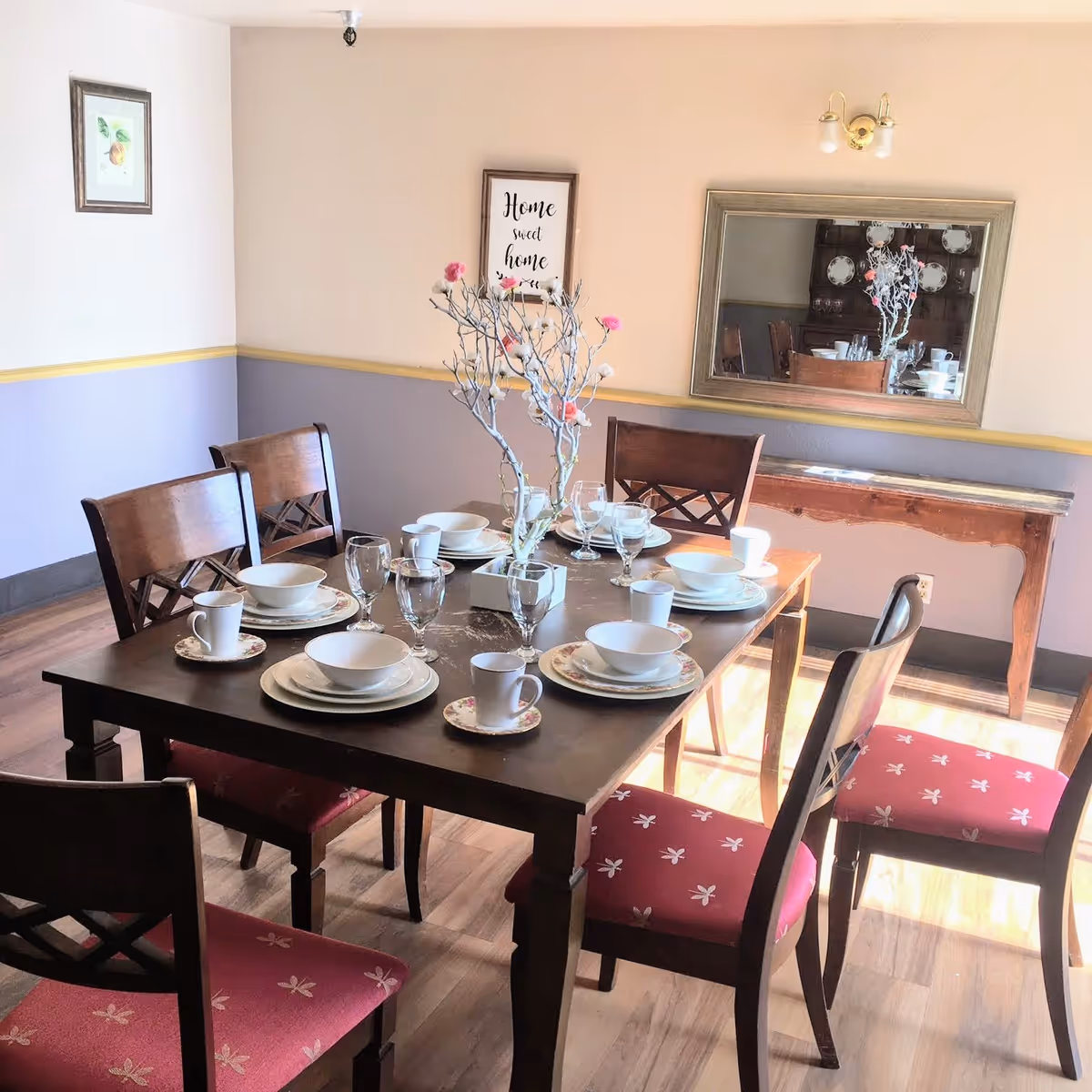 A dining room with a wooden table set for six, floral place settings, a branch centerpiece, and a wall mirror.
