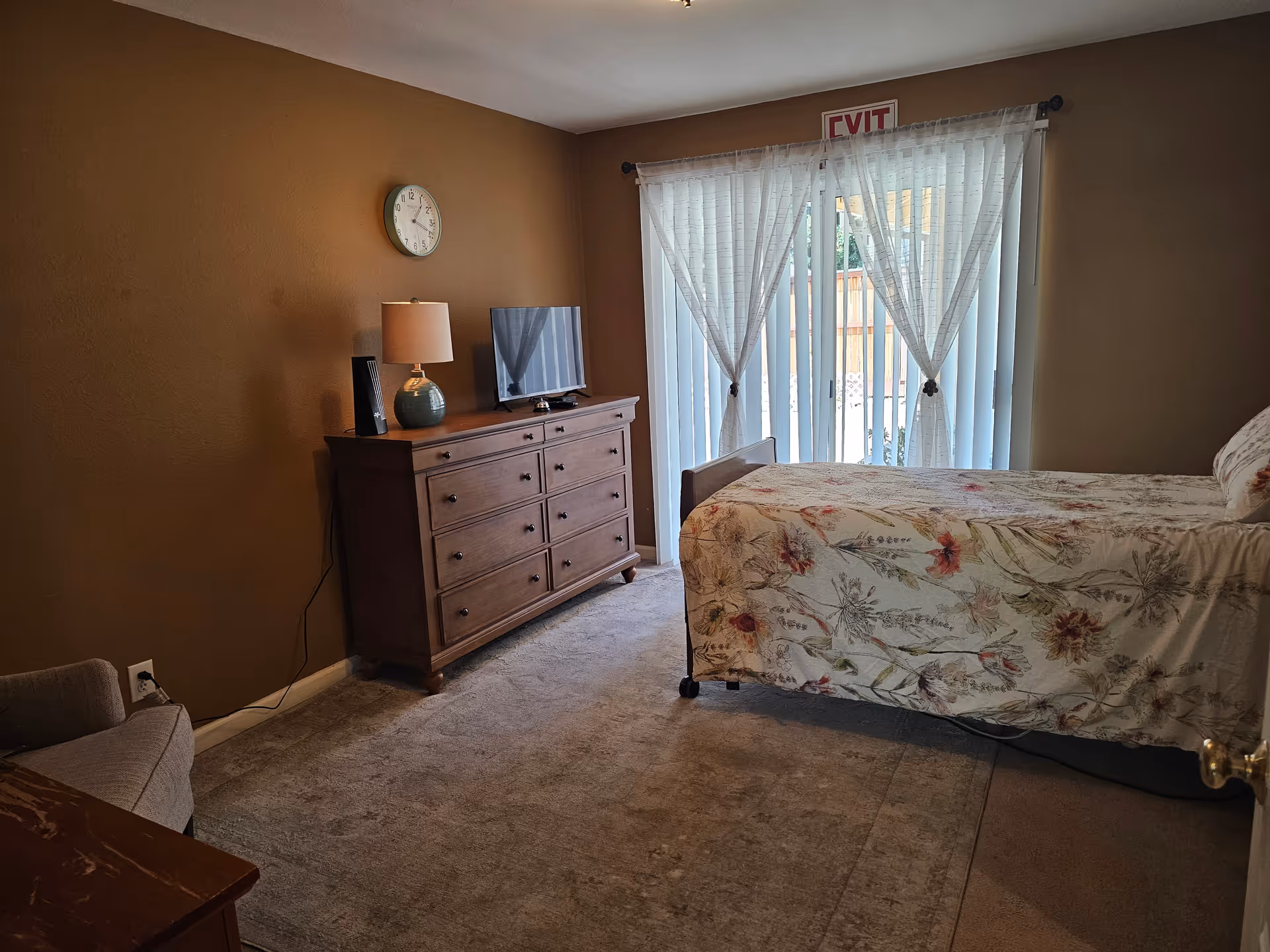 A cozy bedroom with a single bed covered in a floral bedspread, a wooden dresser with a lamp and TV, and sliding glass doors with vertical blinds.
