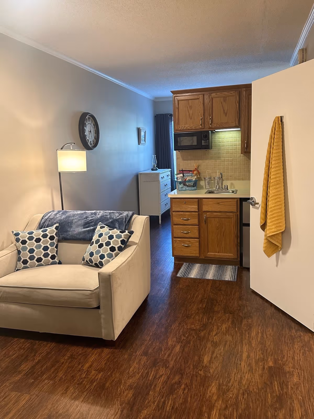 Small studio living area with a beige loveseat, floor lamp, wall clock, and a kitchenette with wooden cabinets and sink.