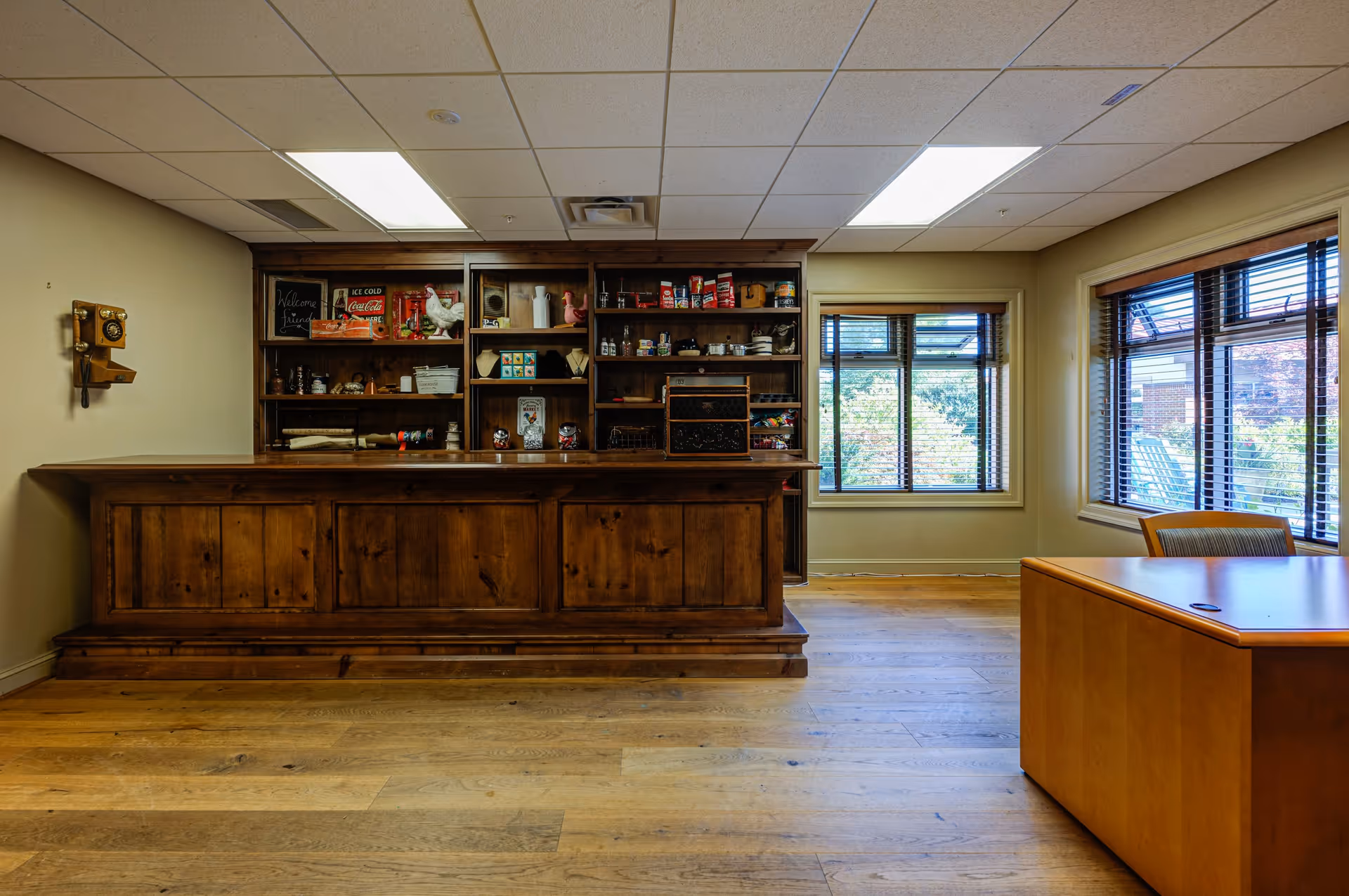Interior room with wooden floor and ceiling tiles, featuring a large wooden counter with shelves behind it containing various decorative items and canned goods. To the right, there is a wooden desk and chair near two windows with blinds, showing greenery outside.