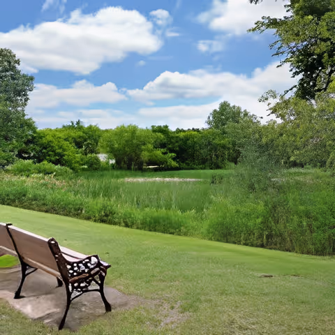 A wooden bench with decorative metal armrests sits on a grassy area facing a lush green landscape with trees and shrubs under a partly cloudy blue sky.