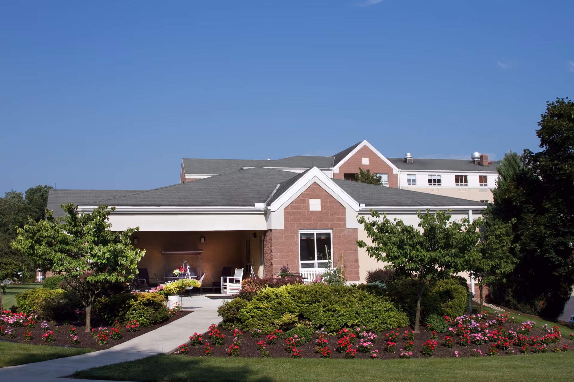 Exterior view of a senior living facility building with a covered porch, surrounded by well-maintained landscaping including bushes, small trees, and colorful flowers under a clear blue sky.