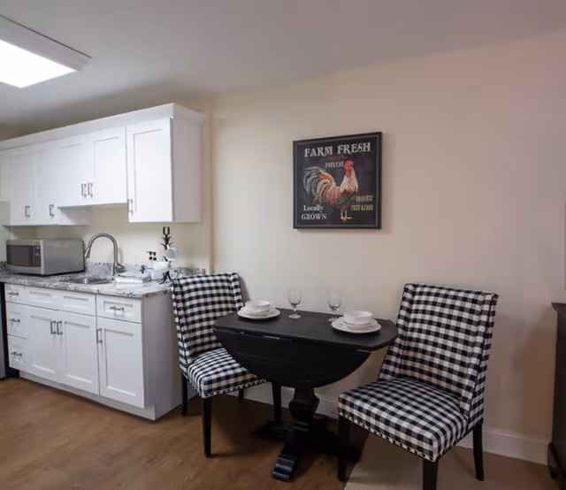 A small kitchen and dining area featuring white cabinets, a granite countertop with a microwave, a sink, and a black drop-leaf table set for two with checkered upholstered chairs. A wall decoration with a rooster and the text 'Farm Fresh' hangs above the table.