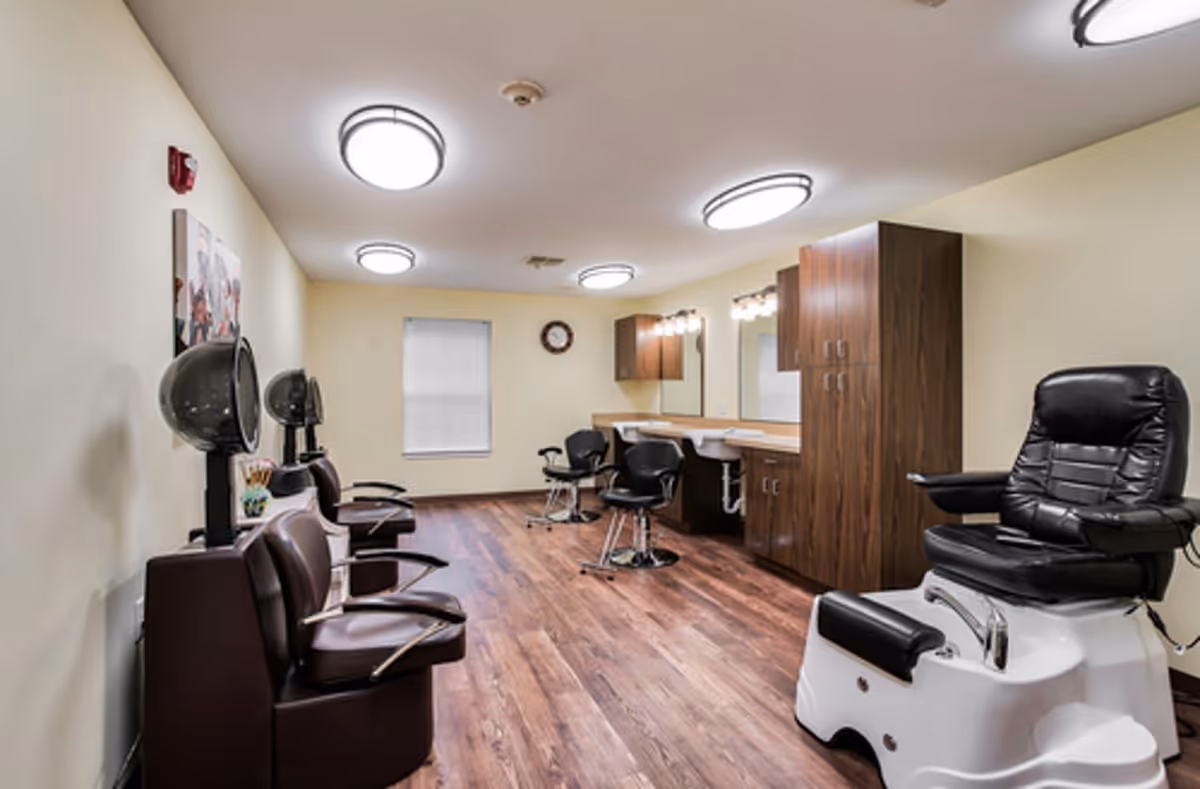Interior view of a salon room in a senior living facility with several salon chairs, hair dryers, a pedicure chair, wooden cabinets, and mirrors along the wall. The room has wood flooring, beige walls, and ceiling lights.