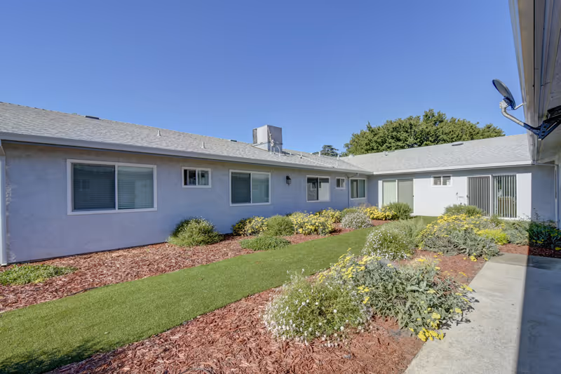 Outdoor courtyard area of a single-story assisted living facility with a well-maintained lawn, flower beds with yellow and white flowers, and a clear blue sky overhead. The building has multiple windows and a sliding glass door.