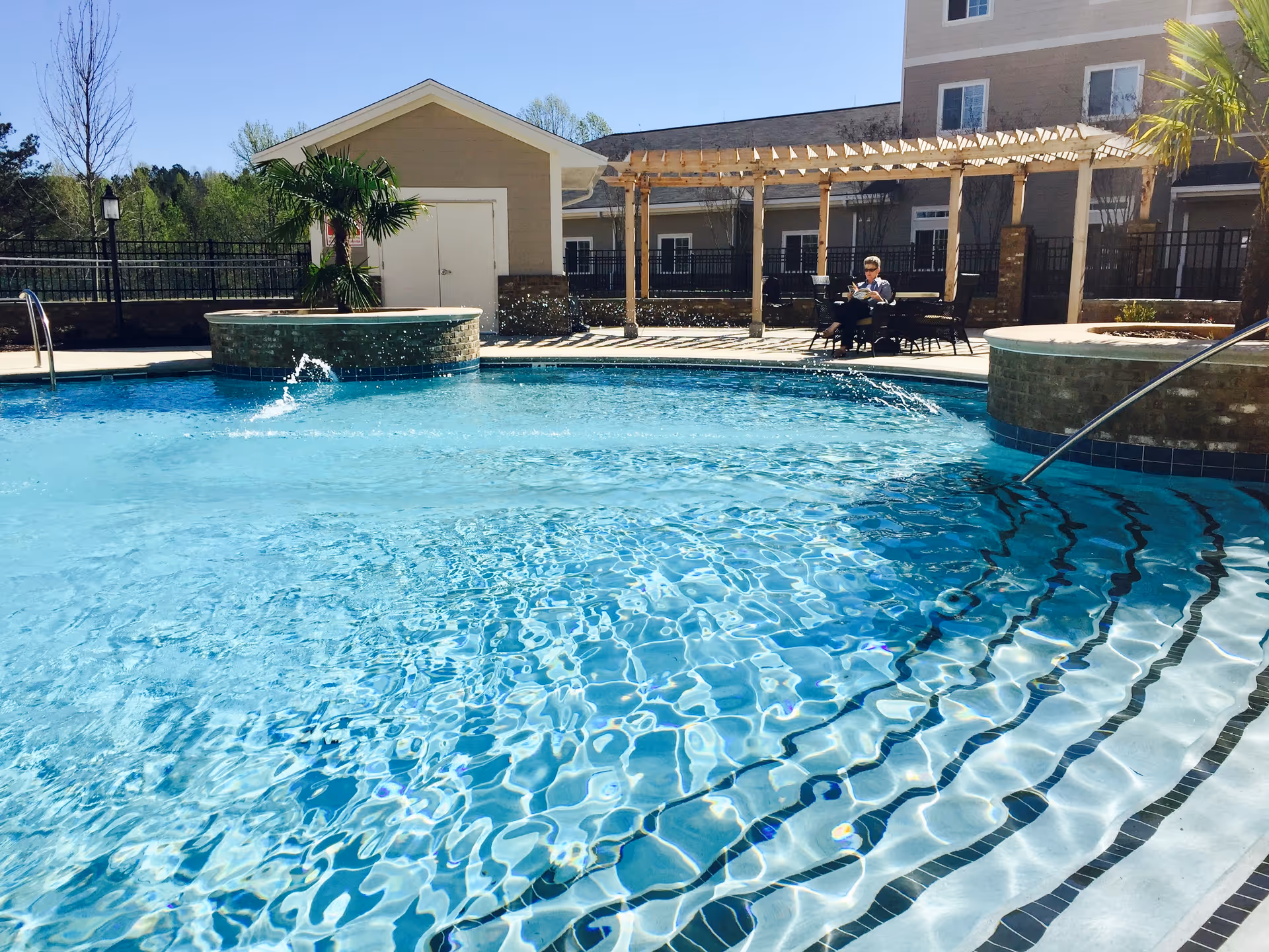 Outdoor swimming pool with clear blue water and water fountains, surrounded by a patio area with chairs and tables. A person is sitting and reading under a wooden pergola. The background shows a building and some trees under a clear blue sky.