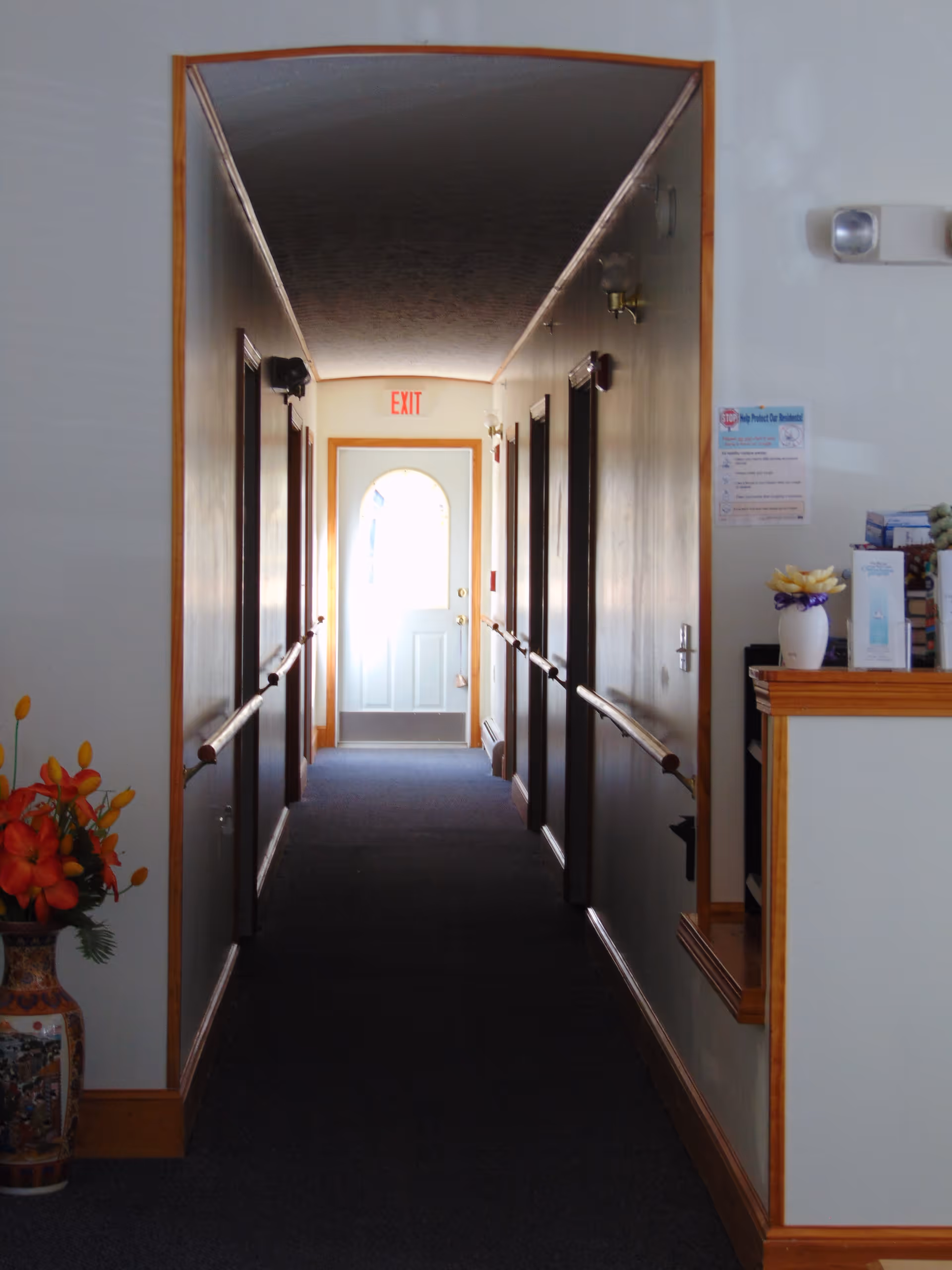 A narrow hallway in an assisted living facility with handrails on both sides, several closed doors along the walls, a vase with orange flowers on the left, and a small reception or counter area on the right. At the end of the hallway is a white door with a window and an illuminated exit sign above it.