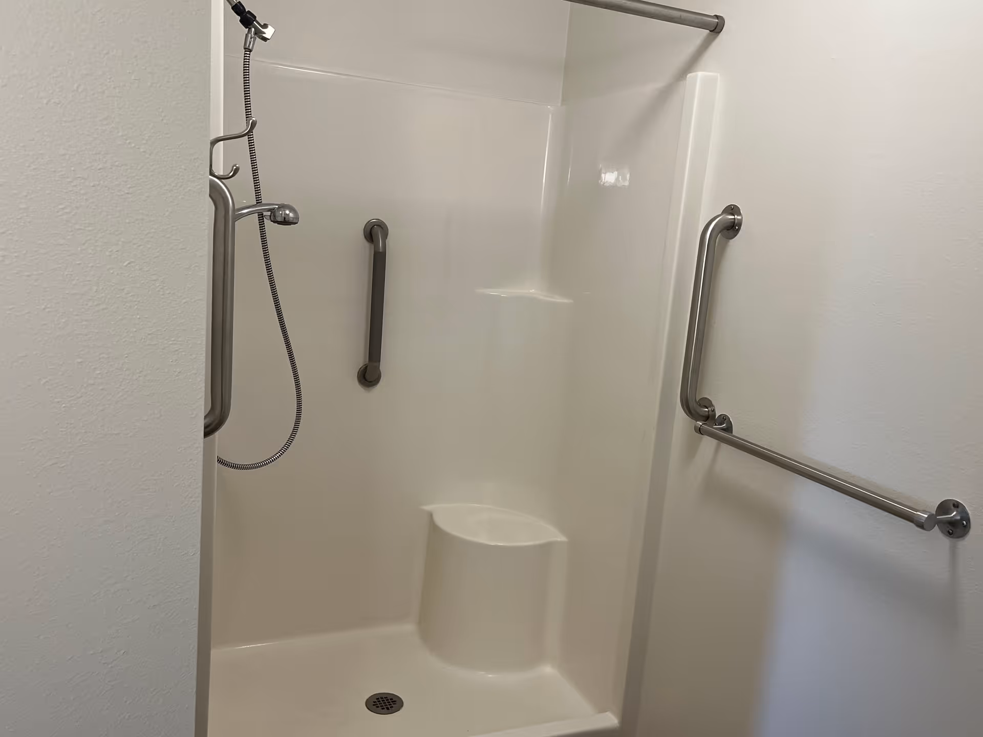 A white shower stall with built-in seat and two stainless steel grab bars on the walls. There is a handheld showerhead attached to a flexible hose and a towel bar mounted on the right wall.