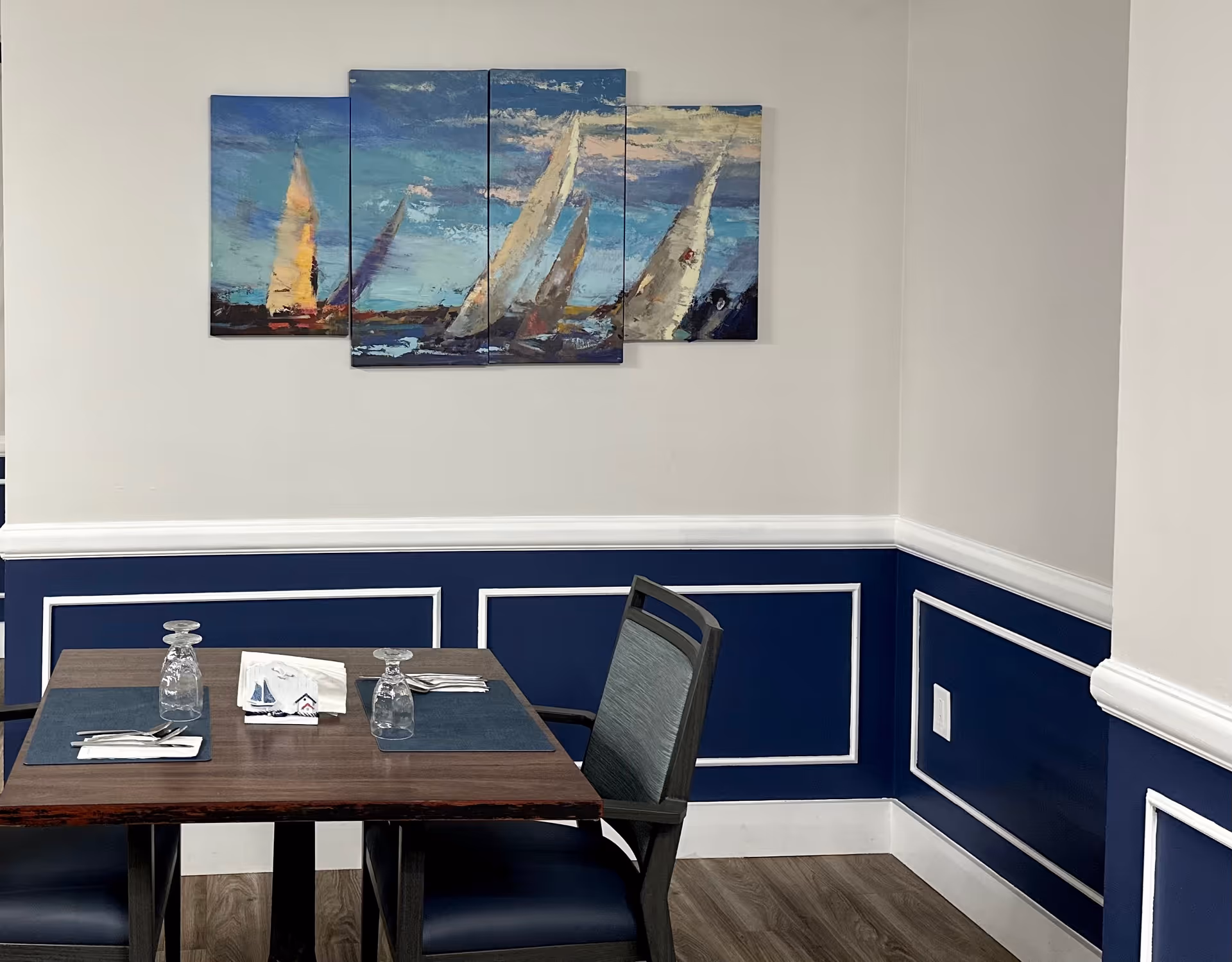 Small dining area with a set table and chairs beneath a multi-panel sailboat painting on white and navy paneled walls.
