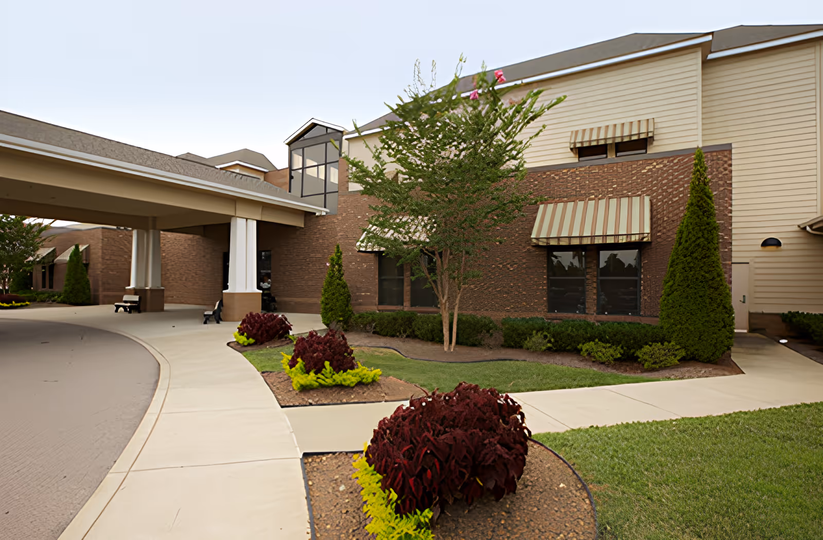 Exterior view of Ahc West Tennessee Transitional Care facility showing a covered entrance with white columns, a curved driveway, landscaped garden beds with red and yellow plants, green shrubs, and a tree. The building has a combination of brick and beige siding with striped awnings over the windows.