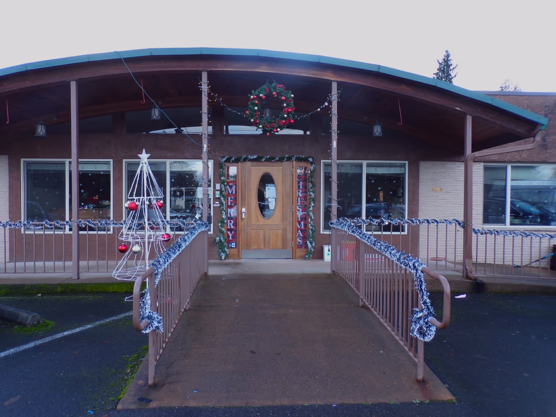Entrance of a building decorated for Christmas with a wooden door framed by garlands and signs reading 'Merry Christmas'. A wreath hangs above the door and a white wireframe Christmas tree with red ornaments is on the left side. The entrance has a covered ramp with railings wrapped in garlands.